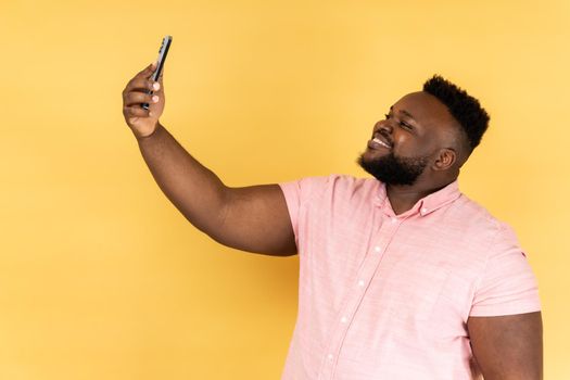 Portrait of positive optimistic man wearing pink shirt talking on video call, having online conversation on mobile phone, taking selfie. Indoor studio shot isolated on yellow background.