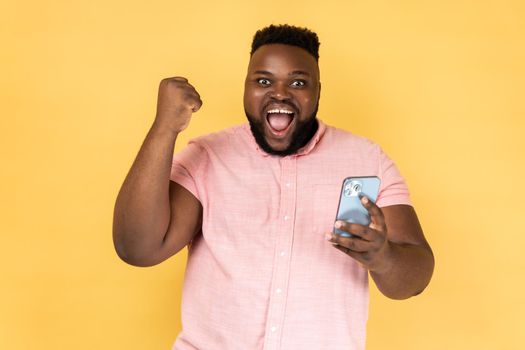 Portrait of satisfied bearded man wearing pink shirt standing with cell phone in hands and clenching fist, celebrating his victory, looks at camera. Indoor studio shot isolated on yellow background.