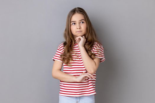 Portrait of thoughtful pensive little dark haired girl wearing striped T-shirt standing holding chin, thinks about something important. Indoor studio shot isolated on gray background.