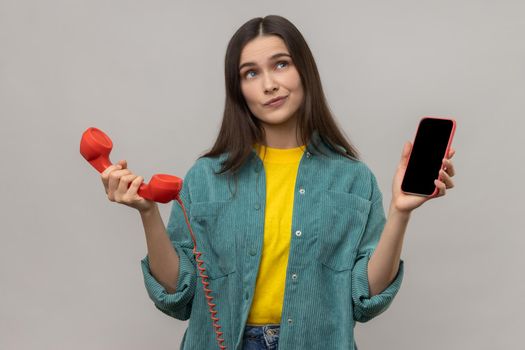 Portrait of confused puzzled woman standing retro telephone and mobile phone, thinking what to use better, wearing casual style jacket. Indoor studio shot isolated on gray background.