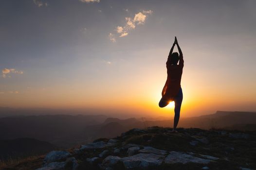 Silhouette of a young woman in a tree pose outdoors in the mountains watching the sunset, yoga balance near the cliff.