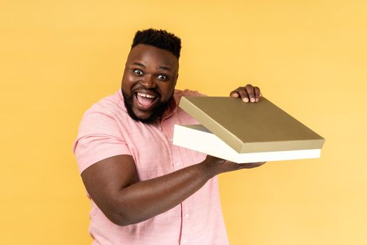 Portrait of cheerful bearded man wearing pink shirt opening gift box and looking at camera with toothy smile, satisfied with present. Indoor studio shot isolated on yellow background.