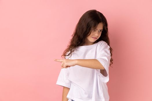 Get out. Portrait of little girl wearing white T-shirt showing exit, demanding to leave her alone, turning away with resentful irritated expression. Indoor studio shot isolated on pink background.
