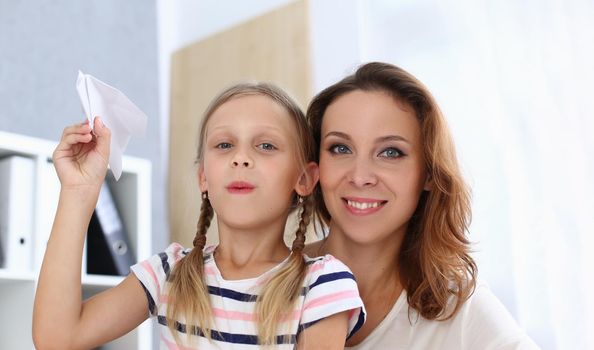 Portrait of beautiful happy mom with daughter are holding paper airplane. Home family pastime and entertainment