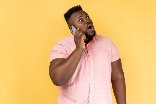 Portrait of shocked man wearing pink shirt talking with friend on cell phone, looking away with surprised amazed, hearing breaking news. Indoor studio shot isolated on yellow background.
