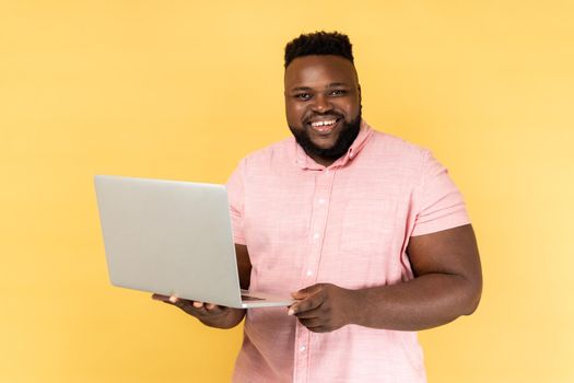 Portrait of smiling freelancer man wearing pink shirt standing and holding laptop, satisfied with teleworking, likes his job, looking at camera. Indoor studio shot isolated on yellow background.