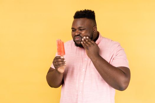 Portrait of unhealthy bearded young adult man wearing pink shirt holding cold ice cream and suffering terrible pain, has sensitive teeth. Indoor studio shot isolated on yellow background.