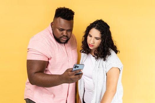 Portrait of serious concentrated young couple in casual clothing standing together, using mobile phone, looking at display, checking social networks. Indoor studio shot isolated on yellow background.
