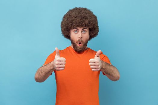 Shocked man with Afro hairstyle wearing orange T-shirt showing thumbs up, looking at camera with open mouth, astonishing with excellent result. Indoor studio shot isolated on blue background.