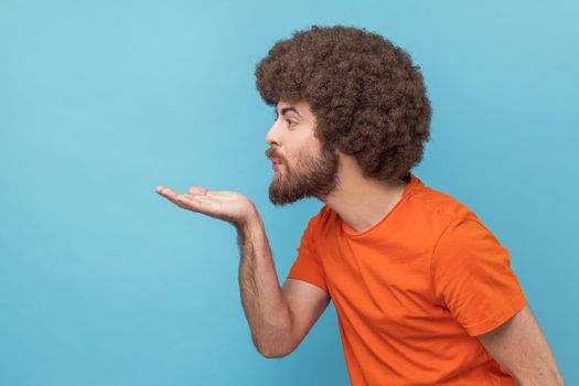 Side view of flirting man with Afro hairstyle wearing orange T-shirt sending romantic air kiss, flirting expressing fondness and affection. Indoor studio shot isolated on blue background.