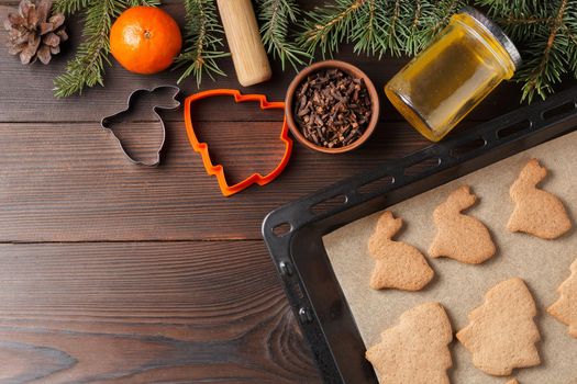 Ready-made Christmas cookies in the form of a rabbit and a Christmas tree on a baking sheet on a wooden table decorated with Christmas tree branches, with a jar of honey and cloves and tangerine. Copy space