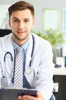 Portrait of a smiling handsome doctor working in clinic. Doctor cardiologist therapist choosing right method of treatment and medical diagnosis sitting at table with patient card