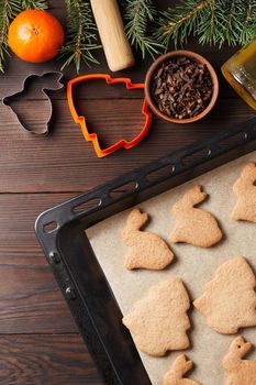 Ready-made Christmas cookies in the form of a rabbit and a Christmas tree on a baking sheet on a wooden table decorated with Christmas tree branches, with a jar of honey and cloves and tangerine.