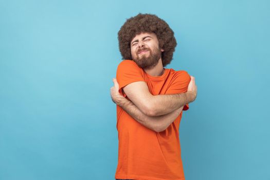 Portrait of selfish narcissistic man with Afro hairstyle embracing himself and smiling with expression of great ego, pleasure and self-esteem. Indoor studio shot isolated on blue background.