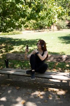 Full body young female athlete browsing cellphone and listening to music in wireless headphones while taking break in running session on sunny summer day in park