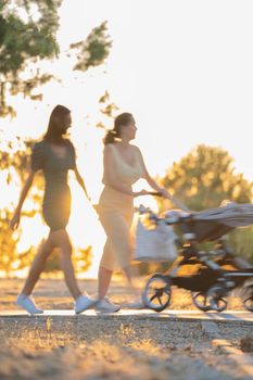 Defocused blurred two girls with a baby cart walking in nature on sunset backlight