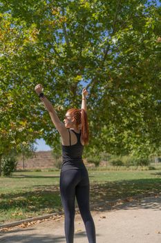 Young redhead female athlete in sportswear raising arms and looking away while celebrating success during fitness training on sunny summer day in park