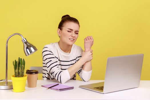 Tired sick woman office manager sitting at workplace with laptop and holding sore wrist, suffering pain in injured hand, carpal tunnel syndrome. Indoor studio studio shot isolated on yellow background