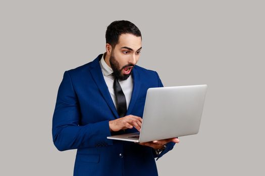 Surprised bearded man holding laptop, typing on keyboard, surfing internet, freelancer working using computer, wearing official style suit. Indoor studio shot isolated on gray background.
