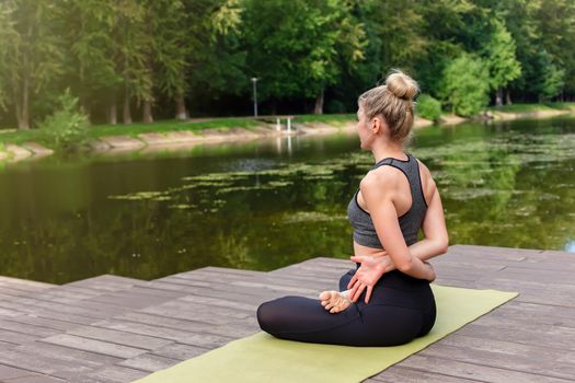 A slender woman in a gray top and leggings, sitting on a wooden platform by a pond in the park in summer and performing yoga. Copy space