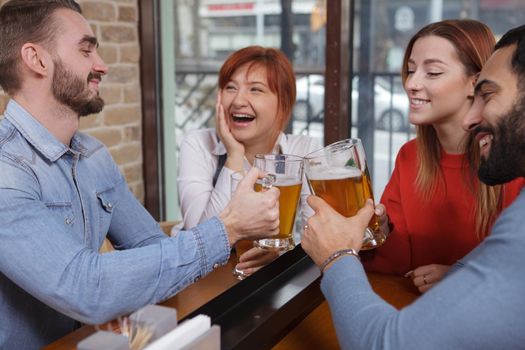 Happy young people laughing, clinking beer glasses at local pub. Cheerful friends drinking beer together