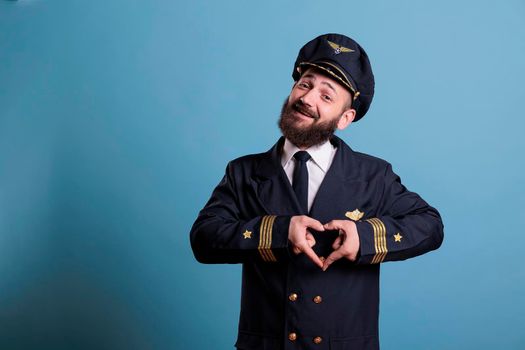 Smiling airplane pilot showing heart shaped love symbol with fingers, conceptual romance gesture. Friendly aircraft captain in uniform expressing affection feelings front view medium shot