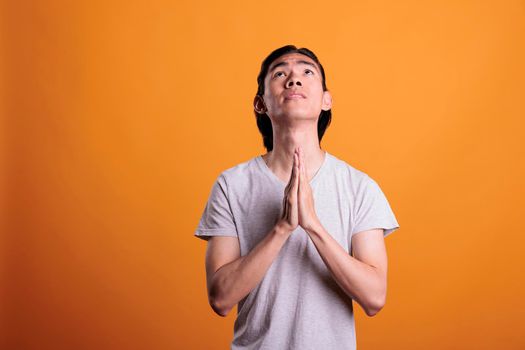 Young man praying folding hands, faith, worship concept. Religious asian teenager pleading, looking upwards with hope, person praising gods, medium shot on orange background