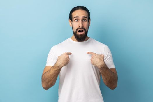 Portrait of amazed happy excited man with beard wearing white T-shirt pointing himself, feeling proud and ambitious, shocked by huge success. Indoor studio shot isolated on blue background.