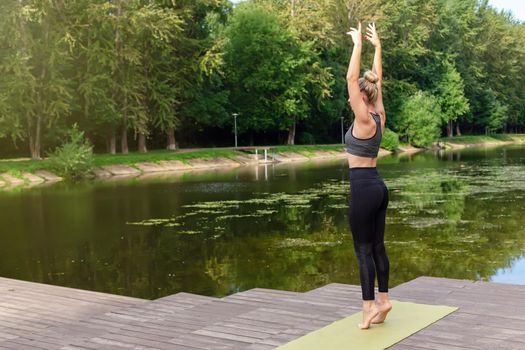 A slender woman stands on a green rug, on a wooden platform by a pond in the park, in summer, with her hands up, doing yoga. Copy space