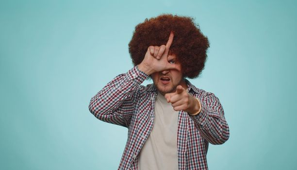 Man showing loser gesture and pointing on you, blaming accusing for unsuccess, expressing disrespect, mocking your failure, bullying abuse. Young guy isolated alone on blue studio wall background