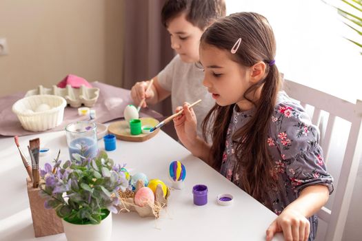 A brunette little girl in gray dress and boy sits at a white table near the window, paints eggs