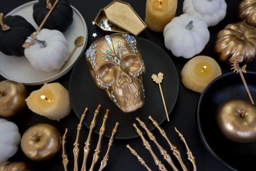 Luxurious table setting for the day of the dead. Luxurious table setting for Halloween. A golden skull with rhinestones lies on a black plate, there are burning candles nearby, there are various pumpkins and golden apples.