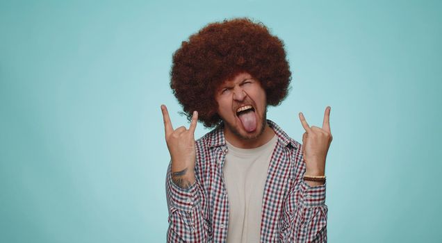Rock n roll. Overjoyed delighted man 30 years old in shirt showing gesture by hands, cool sign, shouting yeah with crazy expression, dancing, emotionally rejoicing in success win alone on blue wall