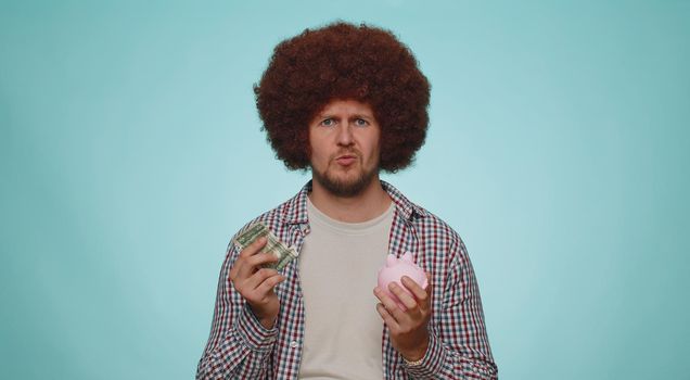 Poor man upset over insufficient amount of money, holding piggybank and one dollar banknote. Financial crisis. Bankruptcy. Poverty and destitution. Young guy isolated on blue studio wall background