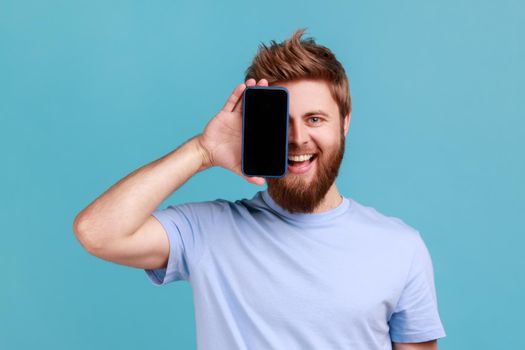 Portrait of smiling optimistic bearded man covering half face with cellphone and looking at camera with toothy smile, positive glad face. Indoor studio shot isolated on blue background.