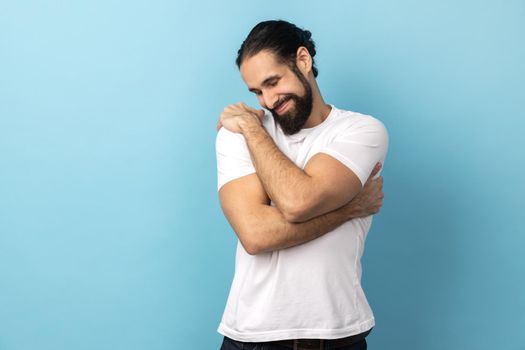 Portrait of happy complacency handsome man with beard wearing white T-shirt standing and hugging himself with closed eyes and toothy smile enjoying. Indoor studio shot isolated on blue background.