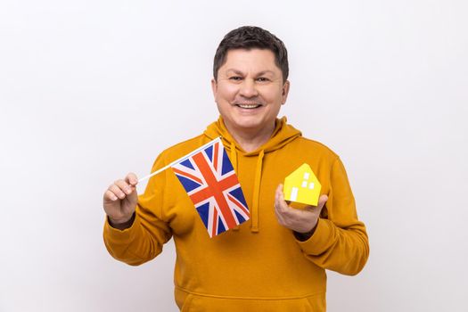 Delighted man holding British flag and paper house, dreaming to buy his own apartrment in Great Britain, looking at camera, wearing urban style hoodie. Indoor studio shot isolated on white background.