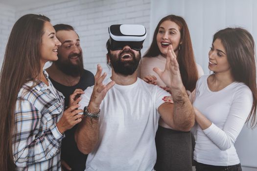 Group of friends surrounding bearded mature man, wearing virtual reality glasses. Team of creative designers using 3d vr goggles at work. Bearded man looking excited, trying virtual reality headset