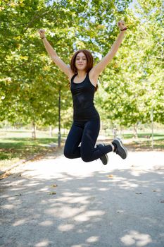 Full body cheerful young female in black clothes raising arms and looking at camera while jumping over asphalt path in sunlit park