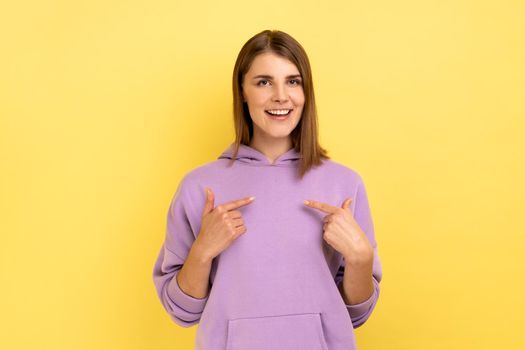 This is me. Attractive woman with dark hair standing with amazed face and pointing herself, being proud and happy, wearing purple hoodie. Indoor studio shot isolated on yellow background.