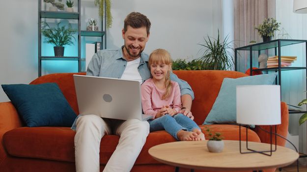 Happy family father or nanny and child kid daughter laugh use laptop, watch cartoons, make video call, look at screen. Dad with child making online shopping on notebook sit on sofa at home living room