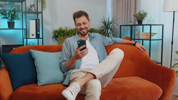 Portrait of adult smiling man sitting on sofa uses mobile phone at modern home apartment. Young guy texting share messages content on smartphone social media applications online, watching relax movie