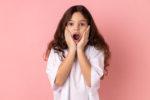 Portrait of amazed little girl wearing white T-shirt standing with hands on cheeks, looking at camera with big eye, saying wow. Indoor studio shot isolated on pink background.