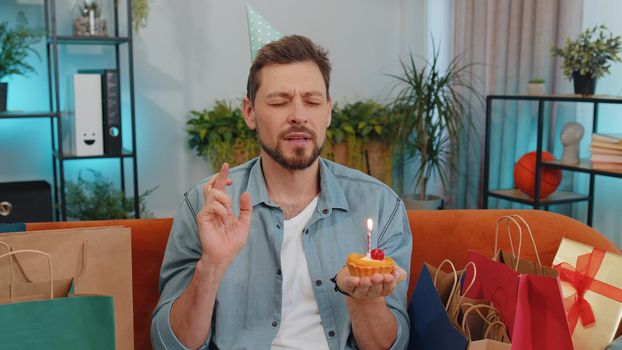 Happy adult man wears festive birthday hat hold cupcake makes wish joyful congratulating blowing burning candle on cake. Young funny guy celebrating anniversary party at home room apartment on couch