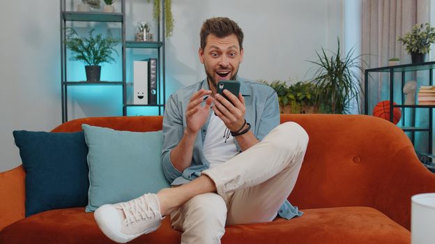 Oh my God Wow. Excited happy joyful winner man use smartphone typing browsing found out great win good news celebrate victory. Young guy with mobile phone sitting at home in room on orange couch