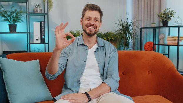 Ok. Happy cheerful handsome man looking approvingly at camera showing ok gesture, positive like sign, approve something good in living room sitting on orange sofa. Young guy at modern home apartment