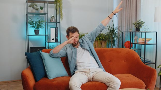 Trendy cheerful positive adult man having fun dancing and moving to rhythm, dabbing raising hands, making dub dance gesture. Young caucasian guy at modern home apartment living room on orange couch