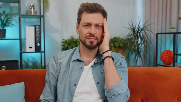 Displeased caucasian adult man rubbing temples to cure headache problem, suffering from tension and migraine, stress, grimacing in pain, high blood pressure. Young guy at home living room on couch
