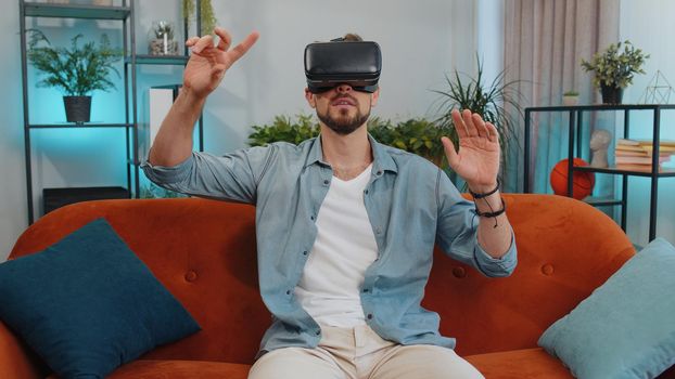 Young man using virtual reality futuristic technology VR app headset helmet to play simulation 3D 360 video game, watching film movie at modern home apartment. Adult guy in goggles sitting on couch