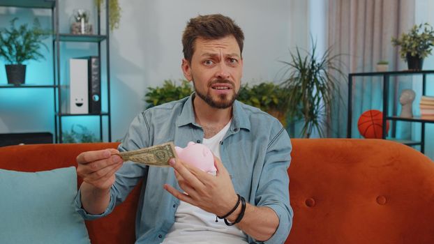 Poor adult man insufficient amount of money, holding piggybank and one dollar banknote at home. Financial crisis. Bankruptcy. Poverty and destitution. Young guy at home apartment living room on couch